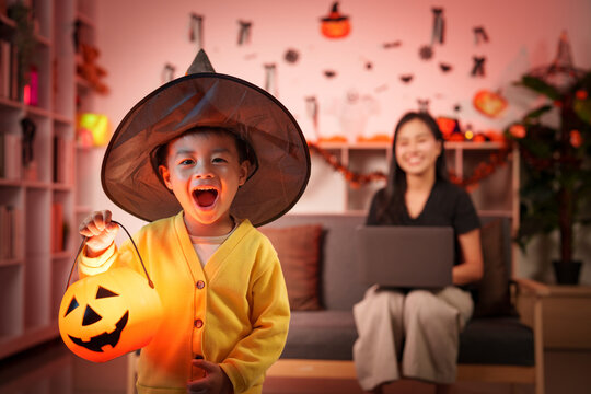 Asian boy in witch hat holding Halloween pumpkin lantern with mother at home