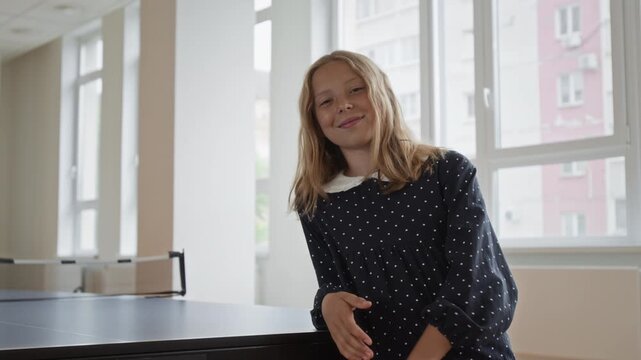 Portrait of Caucasian schoolgirl holding table tennis paddle near ping pong table and smiling at camera in slow motion. Useful for school sports ads, youth education projects, active lifestyle promo