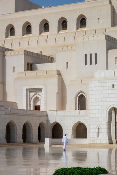 Man in a white dishdasha and cap walking beneath the arched colonnade of the Royal Opera House Muscat facade with latticed windows above, Governorate of Muscat, Muscat, Oman