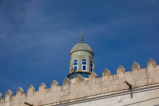 Tiled minaret dome topped with a crescent moon finial rising above a crenellated mosque wall against a clear blue sky, Dhofar Governorate, Wadi Dirba, Oman