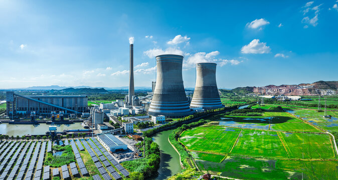 Aerial view of large cooling towers and solar panel arrays next to green fields under bright daylight and blue sky.