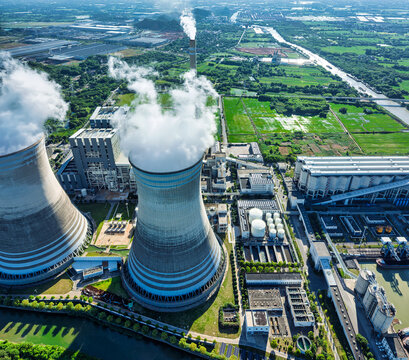 Aerial view of a thermal power plant with cooling towers emitting white steam under bright direct sunlight.