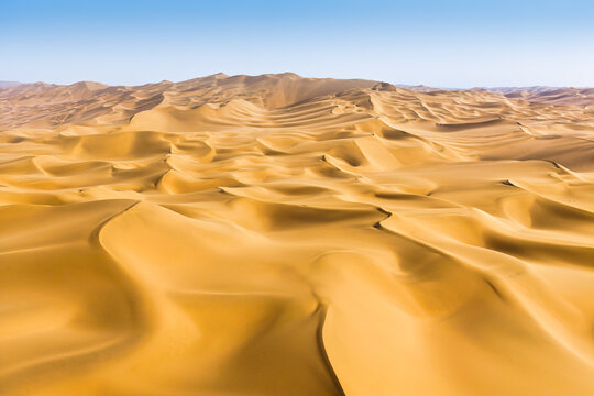 Aerial view of sprawling golden sand dunes under a clear blue sky, showing rolling desert ridges and natural shadow lines under bright daylight.