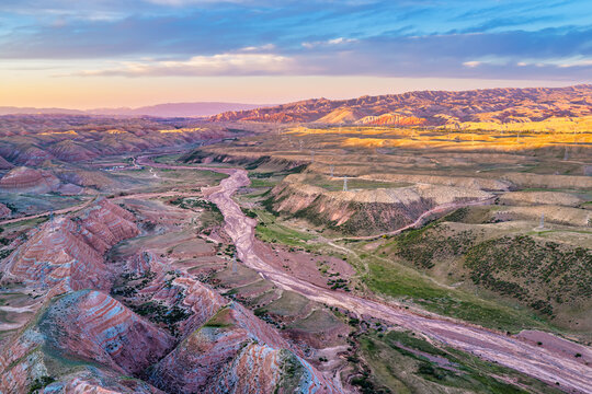 Aerial view of colorful danxia landform rock strata with a dry riverbed and power pylons illuminated by soft sunset light in Xinjiang.