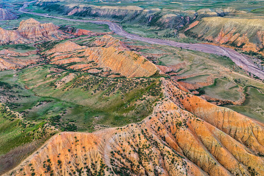 High-angle aerial view of rugged orange sandstone ridges, green grassy slopes, and a dry riverbed under soft natural daylight in Xinjiang.