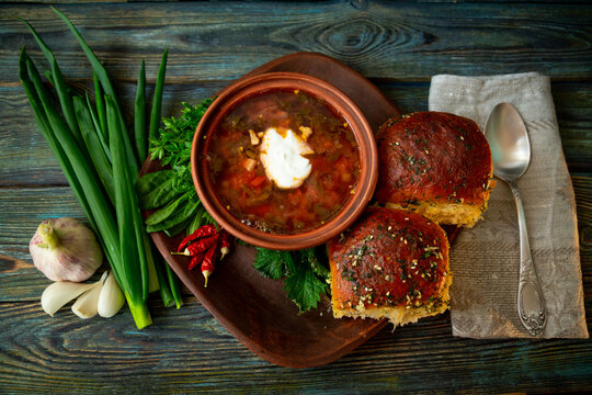 Traditional Ukrainian green borscht soup with nettle, sorrel and sour cream in clay bowl served with garlic pampushky buns, herbs and chili peppers on wooden background