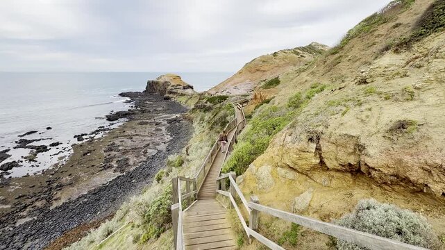 A girl walks on a wooden footpath, admiring the rugged cliff landscape and waves crashing against the rocks at Cape Schanck, Melbourne, Mornington Peninsula, Victoria, Australia.	