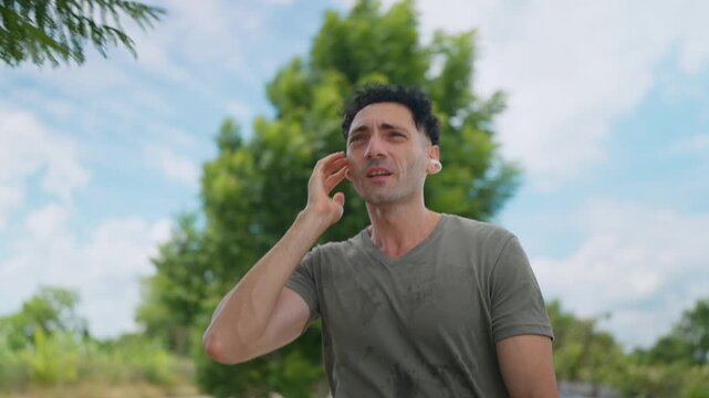 Man stands outdoors, sweating, holding phone to ear. He appears tired, perhaps stressed or frustrated. Trees and sky form a natural, open background