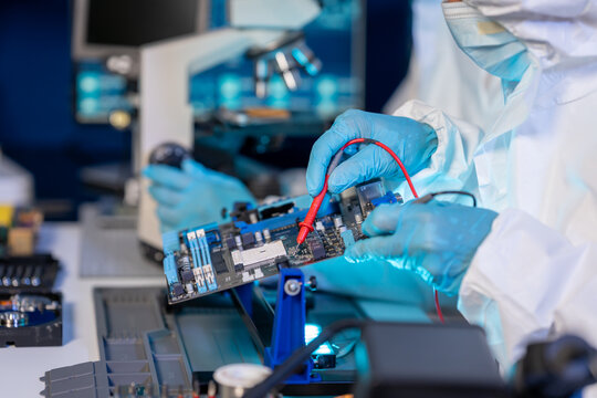 Technician in protective clothing and gloves tests a circuit board with probes in an electronics laboratory, with a microscope and repair tools in the background.