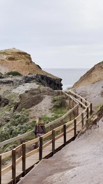 A girl walks on a wooden footpath, admiring the rugged cliff landscape and waves crashing against the rocks at Cape Schanck, Melbourne, Mornington Peninsula, Victoria, Australia.	