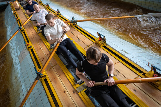 Male rowing crew moves through synchronized stroke cycle beside indoor training pool. Controlled body positions require strength, endurance and concentration despite accumulating fatigue.