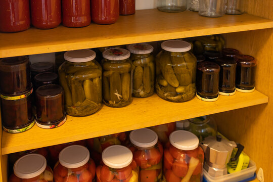 Rows of Homemade Preserved Food in Glass Jars on Wooden Shelves. Several rows of glass jars filled with homemade preserved foods are arranged on wooden shelves. The jars contain different red, yellow,