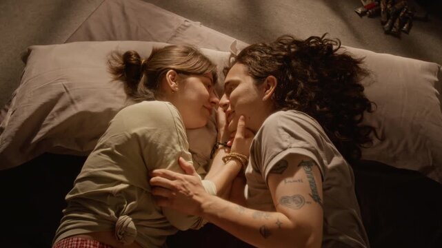Young adult Caucasian couple lying in bed, caressing faces and kissing gently in high angle view. Ideal for romantic campaigns, relationship storytelling and lifestyle branding