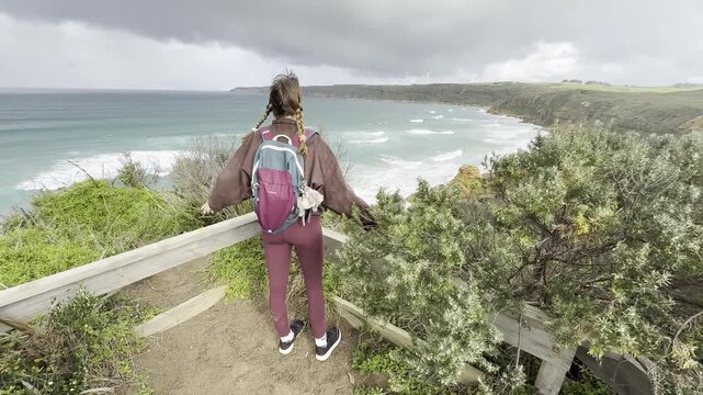 A girl traveler stands on a wooden boardwalk, admiring the stunning rock formations at the beach, Yellow Rock, Portland, Victoria, Australia.
