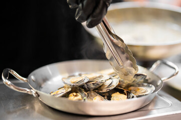 A chef in black gloves uses metal tongs to transfer cooked mussels in a creamy white sauce into a stainless steel serving pan. © pavel siamionov