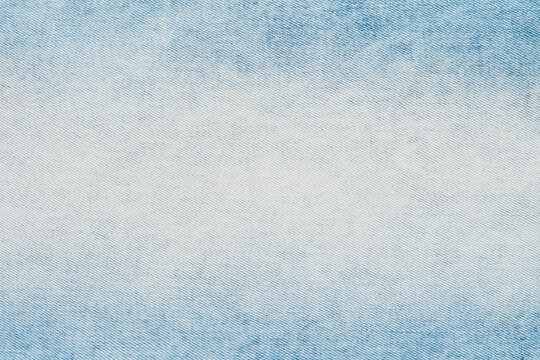 Closeup top view of light blue denim fabric. Stonewashed jeans material shows detailed diagonal weave and faded white center. Neutral, versatile textile background for fashion or design concepts