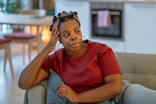 Thoughtful black woman sits at home with hand against head, looking away while feeling stressed, mentally tired. Concept of emotional overload, everyday worries, fatigue, frustration, need for rest.