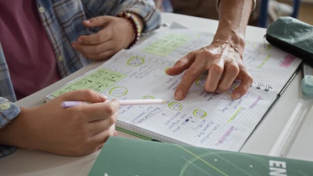 Unrecognizable teacher explaining lesson material to teenage student pointing at notebook with pencil. Ideal for educational platforms, tutoring campaigns, and academic marketing projects