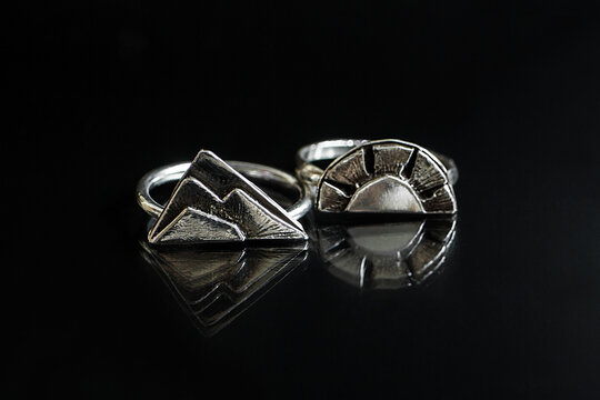 Two silver rings with mountain and sun designs on a reflective black surface. The rings showcase intricate details and a modern aesthetic.