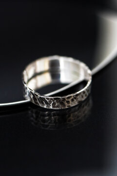 A close-up of a silver ring with a textured surface, resting on a reflective black surface. The ring has a unique design and shines brightly.