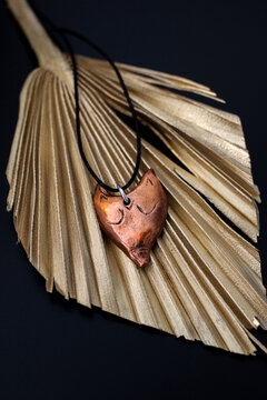 A copper heart pendant hangs on a black cord, resting on a golden palm leaf. The background is dark, highlighting the pendant's intricate design.