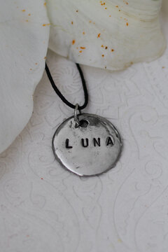 A silver pendant with the word LUNA engraved on it, hanging from a black cord. The pendant is placed on a textured white background with soft lighting.