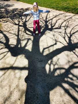 Young girl extends arms wide, playfully aligning herself with the vast, intricate shadow of a tree on a bright park path
