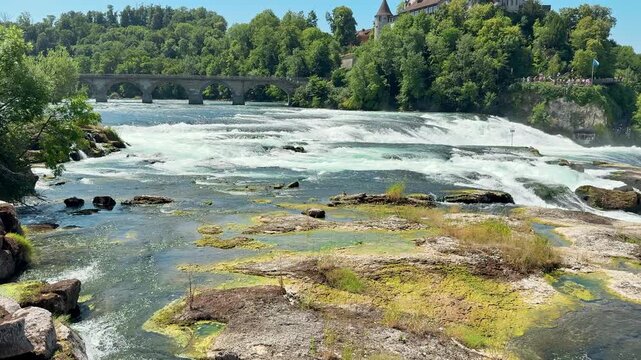 Rhine River flows over wide cascades toward a distant stone bridge outdoors, Calmer foreground water contrasts with white rapids near Schaffhausen