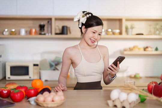 Asian woman using smartphone in kitchen with fresh ingredients for healthy lifestyle