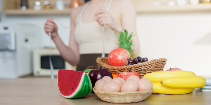 Woman holding tape measure behind fresh fruits and eggs in kitchen for healthy nutrition