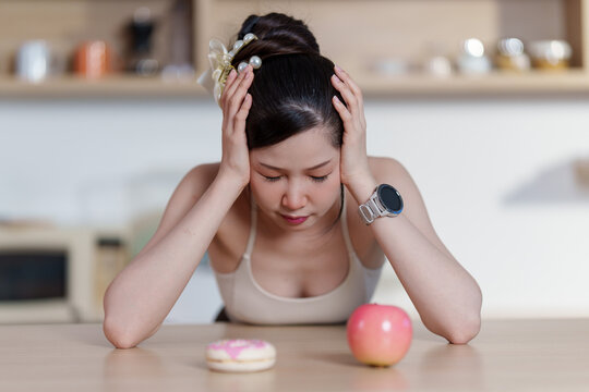 Asian woman struggling with food choice between apple and donut in kitchen