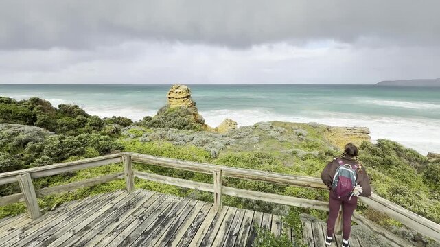 A girl traveler stands on a wooden boardwalk, admiring the stunning rock formations at the beach, Yellow Rock, Portland, Victoria, Australia.