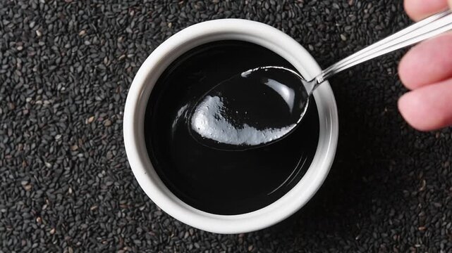 Hand shows thick glossy black sesame paste texture with spoon in ceramic bowl from overhead view.