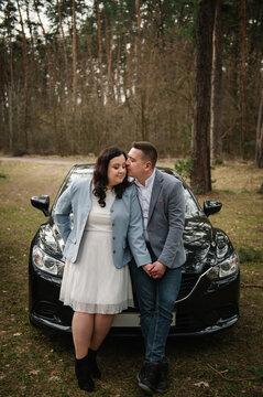 Loving man kissing a smiling woman on the cheek near a black car parked in a pine forest.