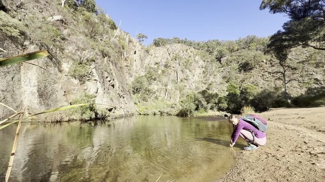 A girl hiker rests by the Werribee River, touching the water, during the challenging Werribee Gorge Circuit Walk - one of the best trails near Melbourne, Victoria, Australia.