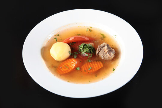 Clear meat broth with vegetables in white plate. Clear meat broth in white bowl. Potato, carrot, tomato, parsley, beef chunks. Healthy homemade soup on dark background. Top view