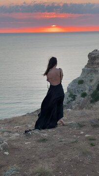 Woman ocean sunset model standing on cliff edge wearing flowing black dress looking at horizon during peaceful evening golden hour travel destination