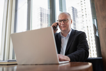 Obraz premium Mature businessman talks on smartphone while using laptop in modern office, representing multitasking, client communication and digital work.