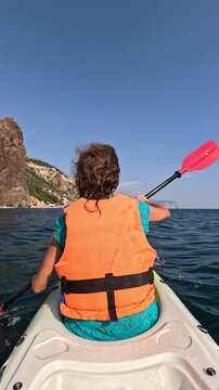 Paddling a kayak through calm open ocean waters past steep coastal cliffs under clear blue skies. Rear view static shot.