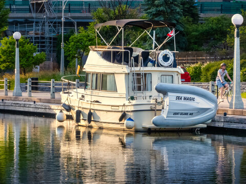Ottawa, Ontario, Canada. Sep 10, 2026. Cabin cruiser boat docked on Rideau Canal near pathway with cyclist riding past in golden light.