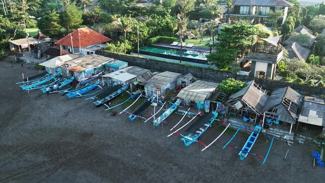 Traditional fishing boats line a black sand beach beside rustic coastal huts and tropical villas with swimming pool creating a strong contrast between local life and tourism push-in aerial footage