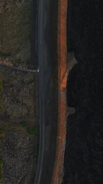 Vertical top-down drone view beside Grindavik, Iceland, showing a dark lava field separated from a paved road by an orange protective berm along the volcanic edge.