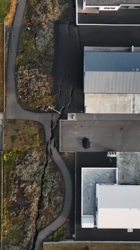 Vertical top-down drone view in Grindavik, Iceland, showing a ground fissure splitting a paved footpath and volcanic terrain between residential buildings after recent unrest.