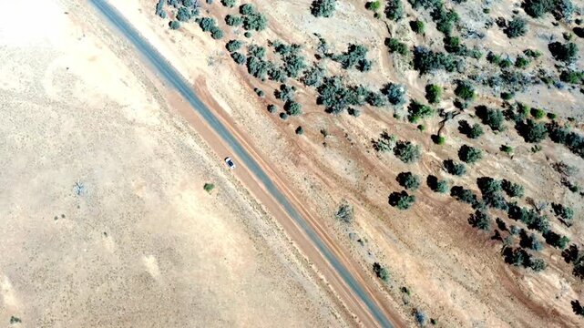 Top-down aerial drone shot tracking a lone car driving a straight outback road, dense native scrub bordering the bitumen amid dry desert plains