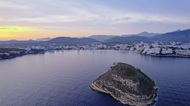 A drone pulls back from the islet of Isla de sa Porrassa, revealing Magaluf's curved bay and hills beyond. Golden sunset light fades into a soft purple-blue evening sky.