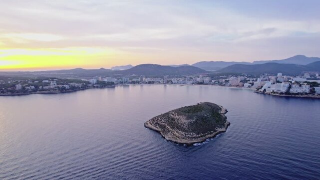 A drone orbits around the small islet of Isla de sa Porrassa, with Magaluf's curved bay and hillside town visible beyond. Golden sunset light fades into a soft blue-violet sky.