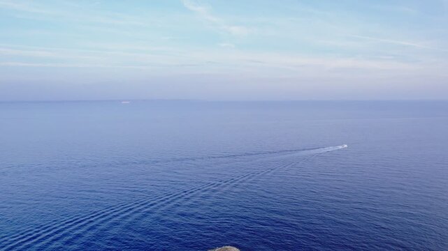 A drone flies forward from the tip of Isla de sa Porrassa while tilting upward, revealing a distant boat crossing calm blue water before the view opens into clear sky and open horizon.