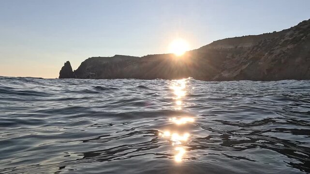 Low angle ocean view of dramatic sunset over rocky coastal cliffs and rippling dark water. First-person handheld video shot.