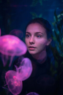 A mesmerized young woman with a fascinated expression looks through thick aquarium glass at beautiful glowing pink jellyfish floating in deep blue water under neon light.