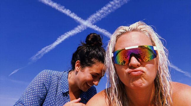Two women making silly faces close up with crossed jet contrails in blue sky
